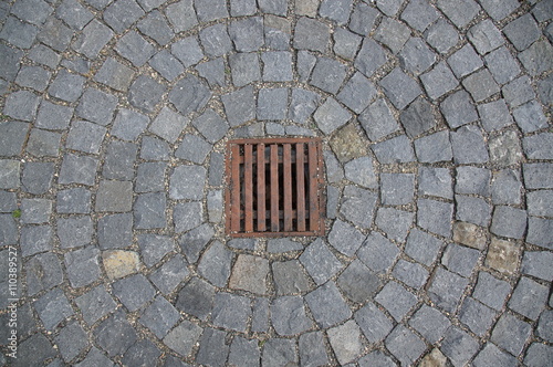 Rostiges Wasserablaufgitter umfasst von Asphaltsteinen von oben fotografiert