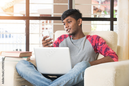 happy looking teenage indian male using laptop and phone