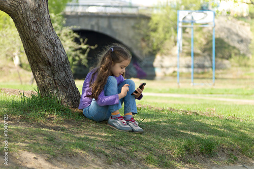 Cute little girl sitting on the grass in a park with a mobile phone in her hands and sending message on phone mobile
