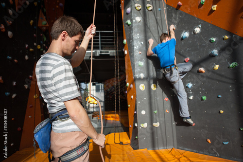Man stands on the ground near rock wall indoors and insuring the climber with rope and carbines