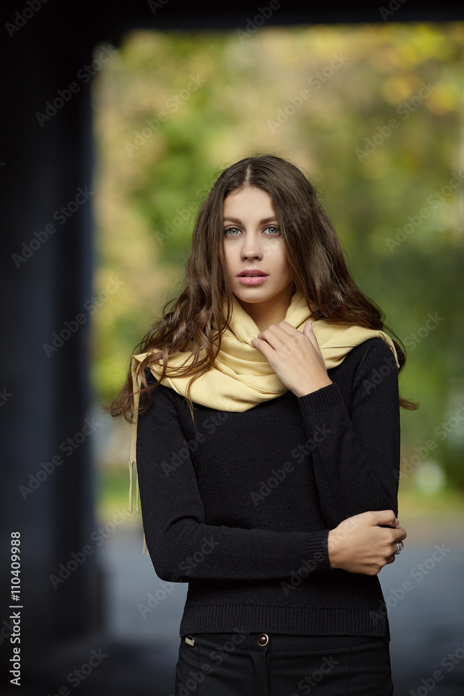 Young romantic brunette woman with long wavy hair posing outdoors in black sweater and yellow scarf with blurry park background Young romantic brunette woman with long wavy hair posing outdoors in black sweater and yellow scarf with blurry park background