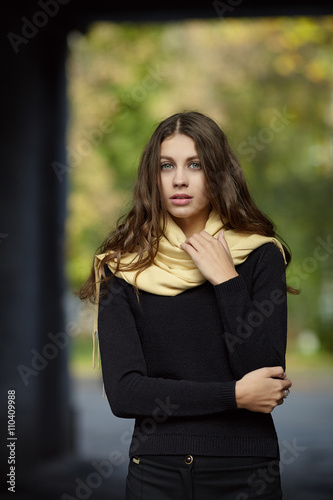 Young romantic brunette woman with long wavy hair posing outdoors in black sweater and yellow scarf with blurry park background