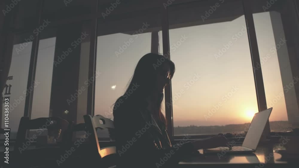 Beautiful young brunette business woman working on her laptop. Heaven sunset glass background behind her. RAW video record.
