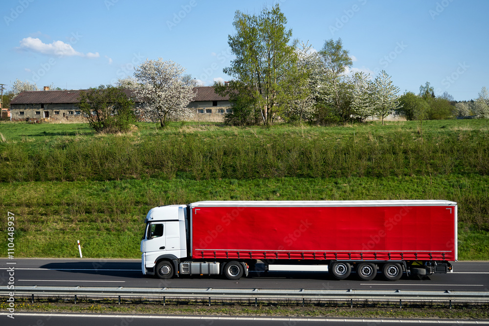 Truck driving on asphalt highway beneath the green grassy slope. Flowering trees, village and old farm building on the horizon. Sunny spring day with blue skies and white clouds.