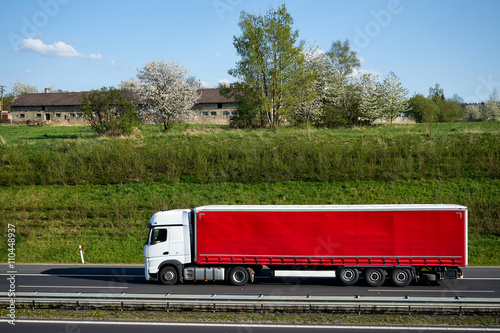 Obraz na płótnie Truck driving on asphalt highway beneath the green grassy slope. Flowering trees, village and old farm building on the horizon. Sunny spring day with blue skies and white clouds. w salonie