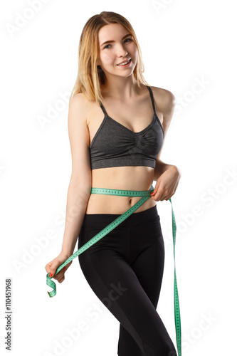 Portrait of attractive cheerful fitness girl smilling and measuring her waist with tape in gray top and black leggings with red dumbbells posing over white background isolated  © garneg