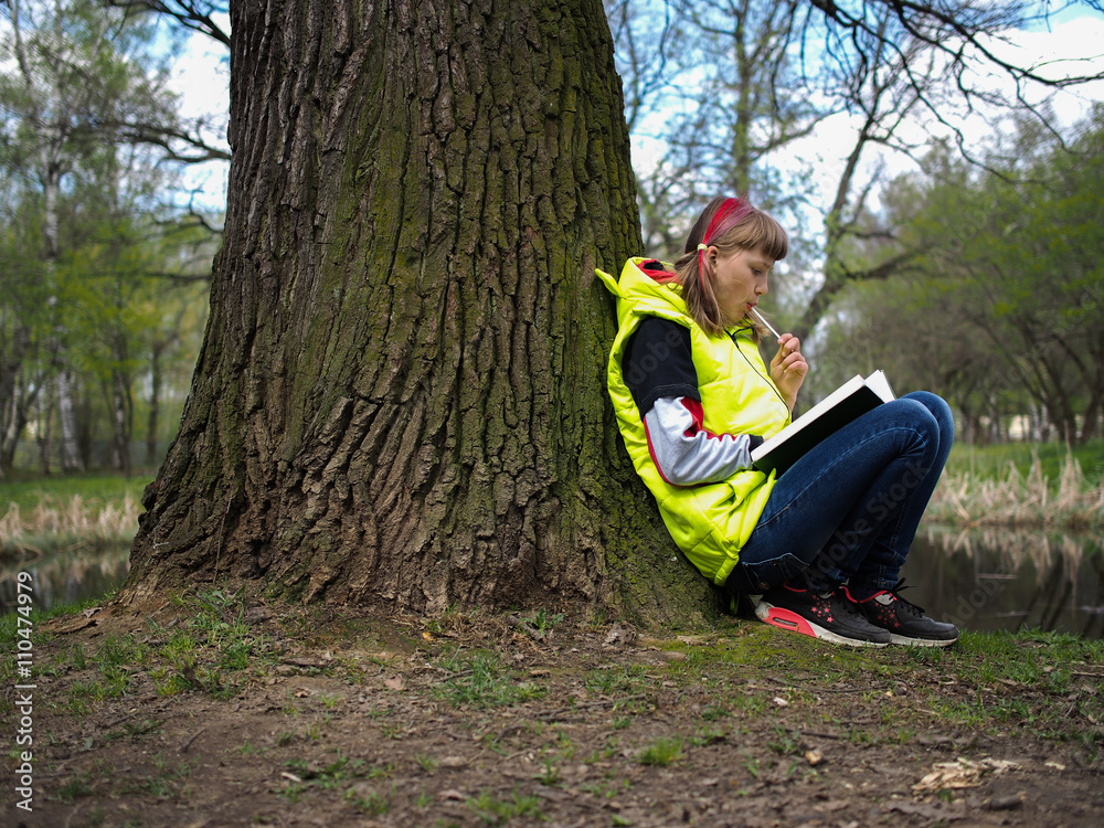 Teen girl in the park with a book. Concept - exam preparation, difficulty learning, school. The girl has her hair dyed red. Bright jacket. Teen girl in the park with a book Teen girl in the park with a book. Concept - exam preparation, difficulty learning, school. The girl has her hair dyed red. Bright jacket. Teen girl in the park with a book