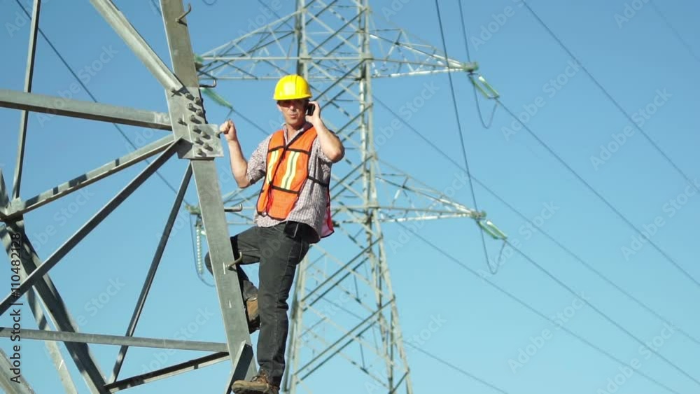 Technician in a safety vest and hard hat standing on the side of a high tension electrical tower and communicates on his cellular mobile phone. Technician in a safety vest and hard hat standing on the side of a high tension electrical tower and communicates on his cellular mobile phone.