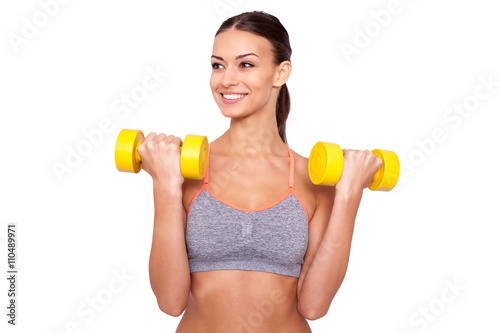 Weight training. Beautiful young sporty woman in tank top exercising with dumbbells and smiling while standing against white background.