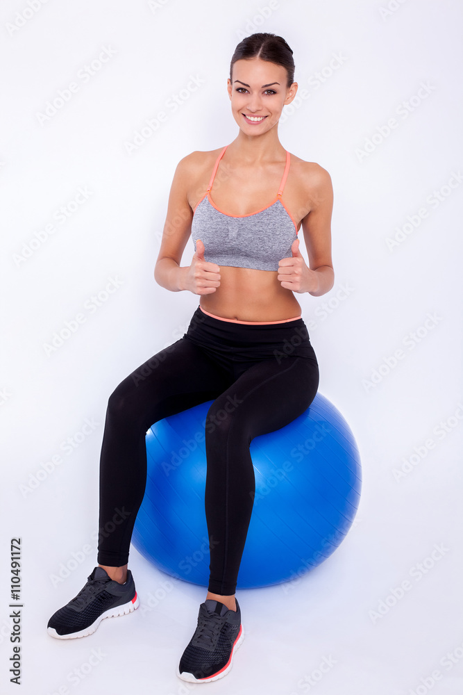 Staying fit. Young beautiful woman in sportswear with perfect bodie thumbs and looking at camera with smile while sitting on fitness balls over white isolated background.