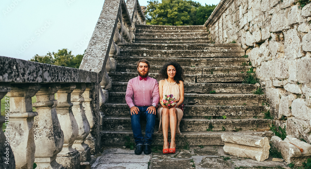 Amazing happy gentle stylish beautiful romantic caucasian couple sits on the ancient stone stairs near the castle. Man with beard, woman with curly hair.