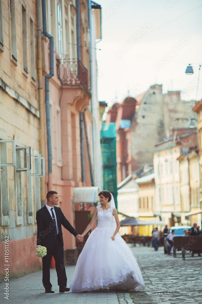 Wedding photo shooting. Bride and bridegroom walking in the city. Married couple embracing and looking at each other. Holding bouquet. Outdoor, full body