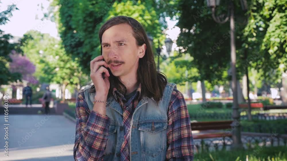Young hipster man with mustache and long hair talking on phone in park Young hipster man with mustache and long hair talking on phone in park