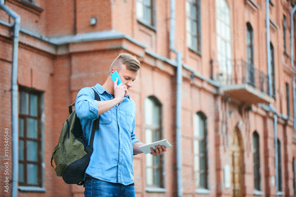 Fototapeta premium Student in a denim shirt, talking on a cell phone and holds your tablet in hand on the background of the building of brick