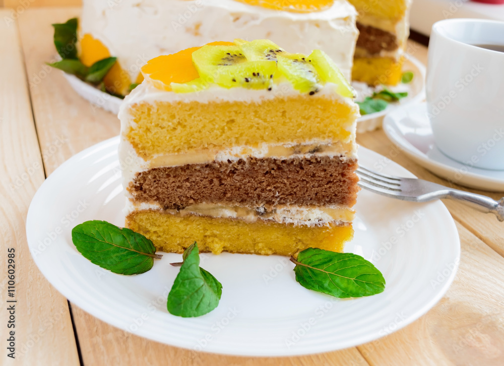 A piece of fruit cake (kiwi, orange, mint leaves) on a white plate on wooden background close-up and a cup of tea A piece of fruit cake (kiwi, orange, mint leaves) on a white plate on wooden background close-up and a cup of tea