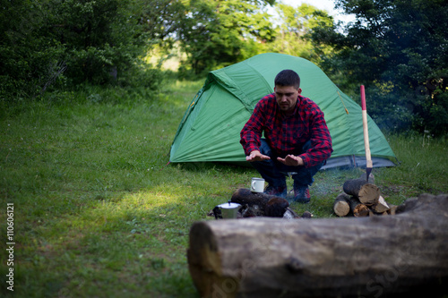 Man drinking coffee by the fire at a campsite on the river bank.