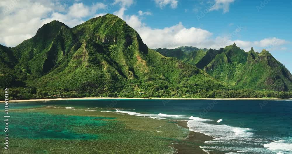 Aerial view flying over tropical coral reef and breaking ocean waves towards beautiful green mountains on Kauai