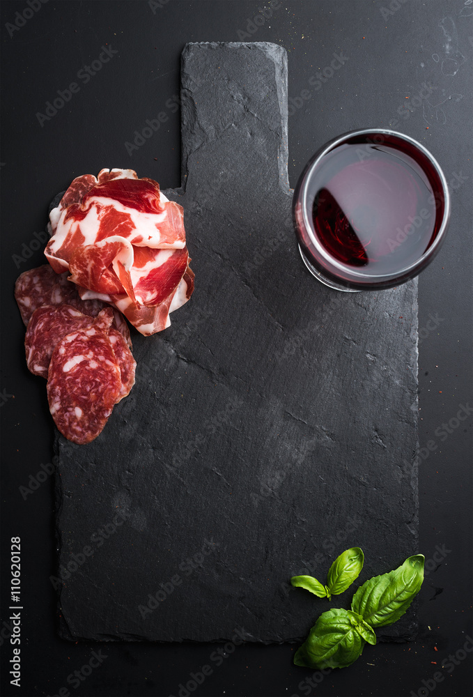 Glass of red wine, meat appetizer and basil on black  slate stone board over dark background