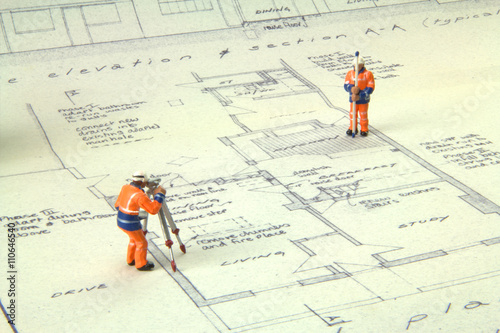 Two miniature figures in high-vis uniform with a theodolite survey a house blueprint photo