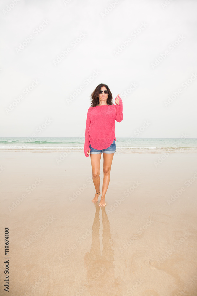 brunette summer vacation woman with sunglasses, red sweater blue jeans shorts barefoot standing finger and hand up on sand with ocean behind, in beach in Cadiz, Andalusia, Spain, Europe