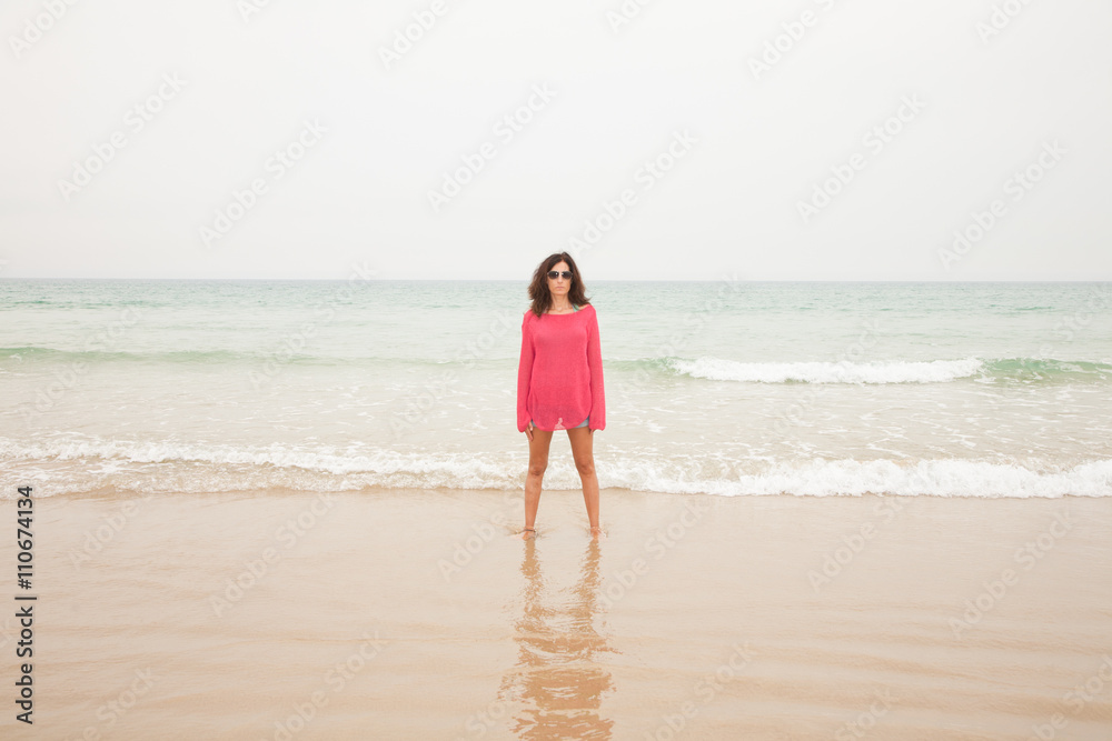 brunette summer vacation woman with sunglasses, red sweater blue jeans shorts barefoot standing at seashore on sand with ocean behind, in beach in Cadiz, Andalusia, Spain, Europe
