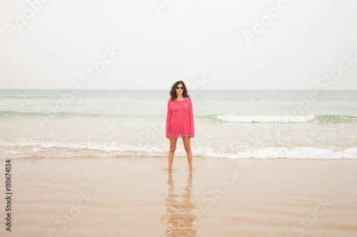 brunette summer vacation woman with sunglasses, red sweater blue jeans shorts barefoot standing at seashore on sand with ocean behind, in beach in Cadiz, Andalusia, Spain, Europe 