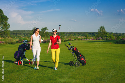 Two smiling sportive women golfers walking on golf course at sunny day