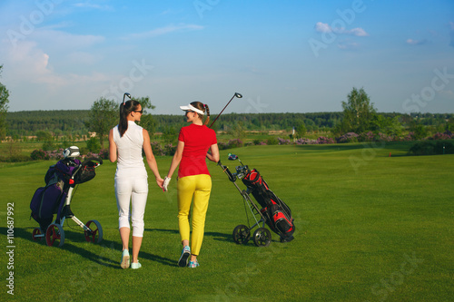 Two smiling sportive women golfers walking on golf course at sunny day
