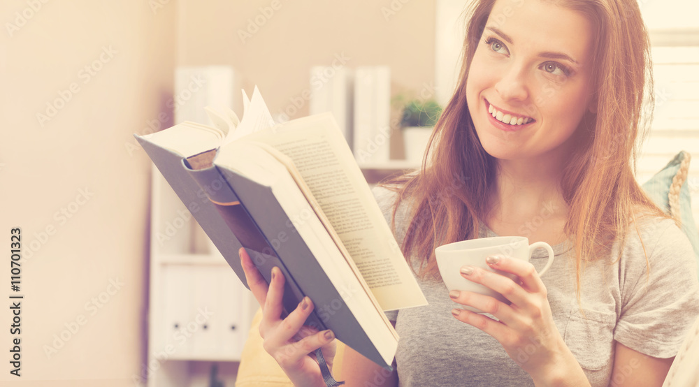 Happy young woman reading a book and drinking coffee on her couch Happy young woman reading a book and drinking coffee on her couch