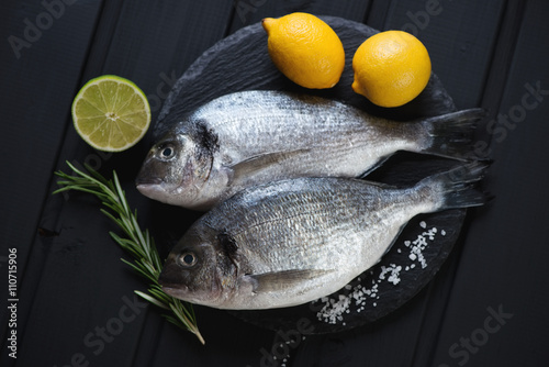 Stone slate with raw dorados on a black wooden surface, top view