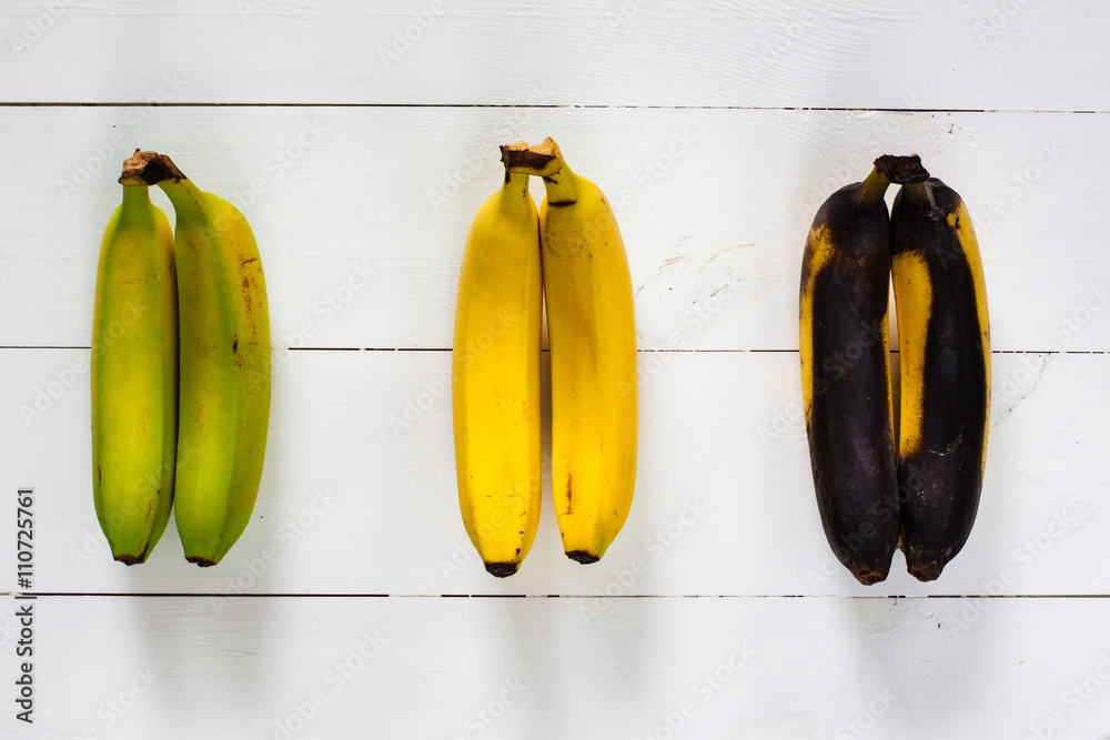 Green, yellow and black bananas arranged in a row on white wooden table.