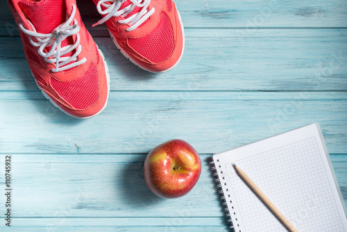 Fitness concept, pink sneakers, red apple and notebook with pencil on wooden background, top view