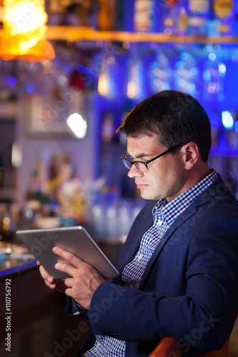 Businessman wearing glasses sitting at a table reading a tablet in a brightly lit colorful cocktail bar, close up side view