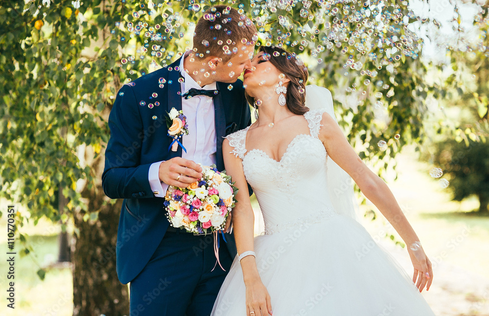 elegant stylish groom with his happy gorgeous blonde bride have fun with soap bubble on the background of trees outdoors in park