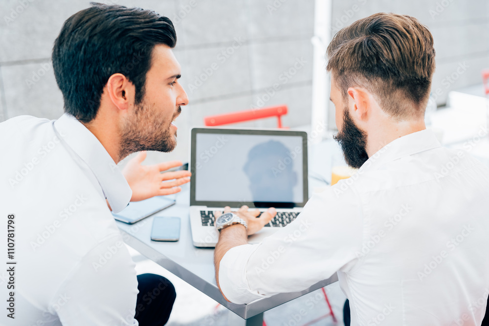 Rear view of two young bearded caucasian modern business man sitting in a bar, using smart phone and laptop, looking downward the screen, smiling - business, work, technology concept Rear view of two young bearded caucasian modern business man sitting in a bar, using smart phone and laptop, looking downward the screen, smiling - business, work, technology concept