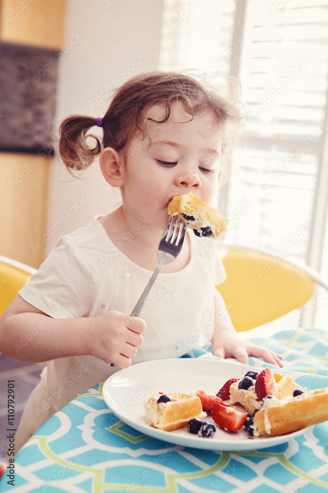 Portrait of one happy white Caucasian kid girl toddler with pig-tails in white dress eating breakfast waffles fruits with fork in sunny kitchen early morning Portrait of one happy white Caucasian kid girl toddler with pig-tails in white dress eating breakfast waffles fruits with fork in sunny kitchen early morning