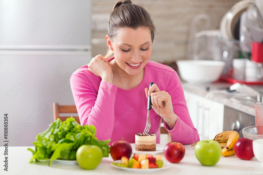 Young woman is eating a cake instead of healthy fresh fruit and vegetable on the table Young woman is eating a cake instead of healthy fresh fruit and vegetable on the table