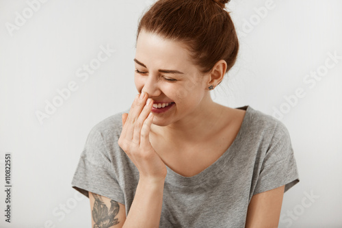 Vivacious teenage girl giglling with her eyes screwed up in a moment of fun as she sitting against white studio wall. Happy natural laughing young casual female covering mouth. Human face expressions