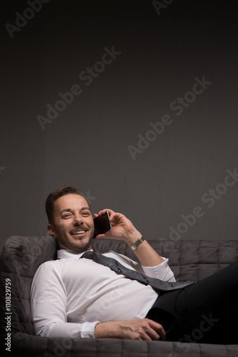 Portrait of handsome businessman talking over mobile or smart phone while lying on comfortable sofa in office.