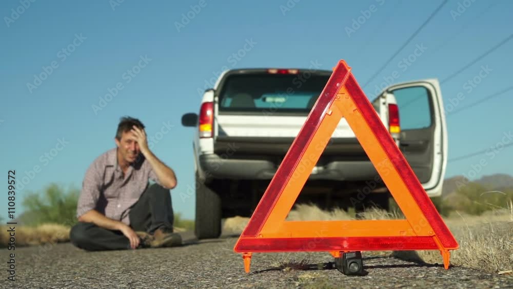 Male in the distance sits on the roadside with his broken down truck with hazard lights on parked on the side of the road and an emergency marker reflective triangle in the foreground.