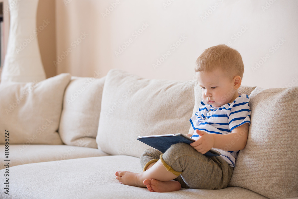 Adorable blond toddler boy laying on the sofa and playing with tablet pc at home, indoors. Child with tablet computer. Adorable blond toddler boy laying on the sofa and playing with tablet pc at home, indoors. Child with tablet computer.