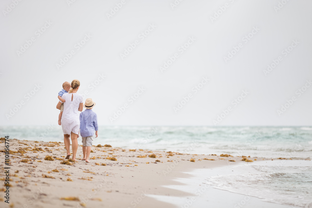 Young mother in white dress walking with her two little boy along the ocean beach. Woman with a baby and a boy enjoying vacation by the sea. On the empty beach. Motherhood.