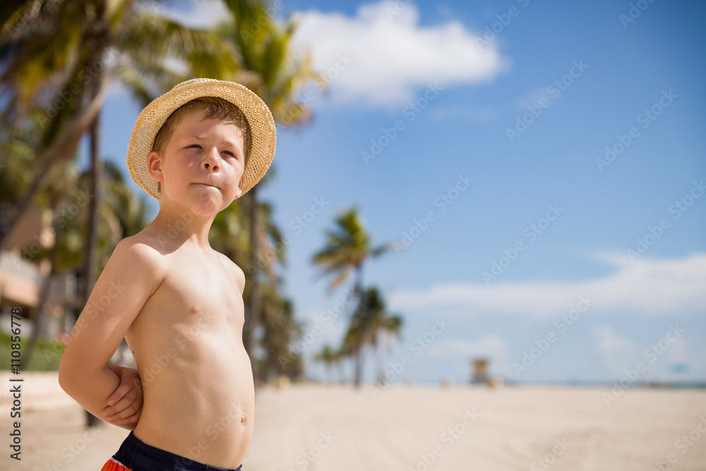 Portrait of adorable kid boy in a straw hat standing on the sandy beach,,  looking into