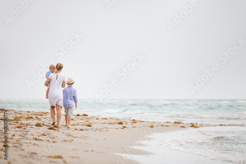 Young mother in white dress walking with her two little boy along the ocean beach. Woman with a baby and a boy enjoying vacation by the sea. On the empty beach. Motherhood.