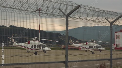 Wallpaper Mural Two helicopters standing on  helicopter pad or platform against background with sunset, mountain covered with forests and metal fence or barbed wire in the foreground.Rack focus, wide shot.  Torontodigital.ca