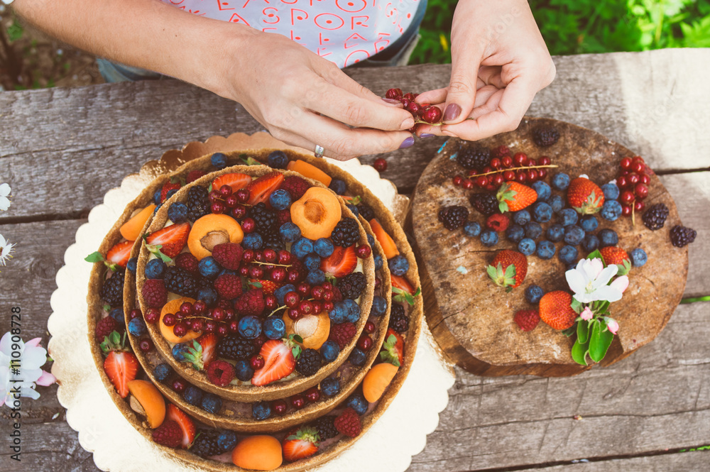 Woman hands of baker carefully decorate cheesecake with fruits and berries on the old planks, white apple and cherry flowers