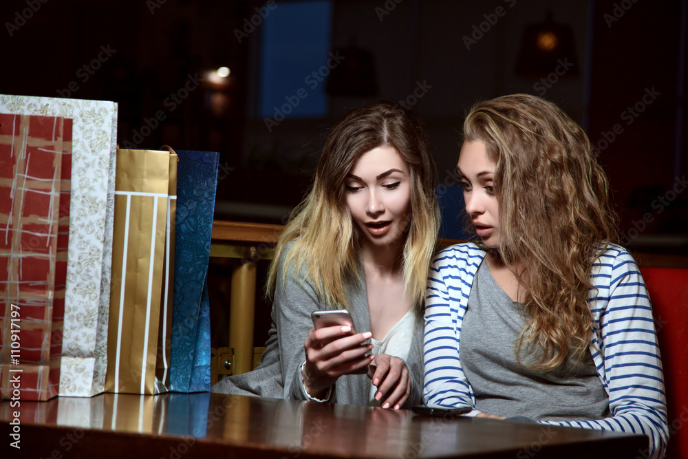 Beautiful girls sitting at a table in a cafe and looking at mobile phone. Fashion Shopping two ladies Portrait. Beauty Woman with Shopping Bags in Shopping Mall. Shopper. Beautiful girls sitting at a table in a cafe and looking at mobile phone. Fashion Shopping two ladies Portrait. Beauty Woman with Shopping Bags in Shopping Mall. Shopper.