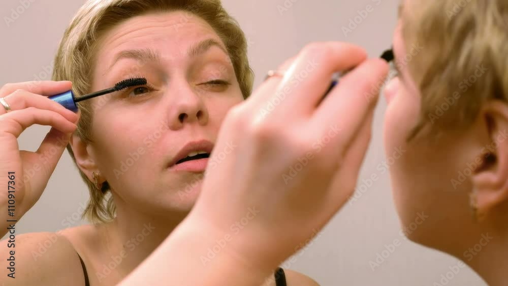 Pretty blond woman applying mascara make-up on her eyelashes in front of mirror and looks at herself. Beauty and makeup concept. 4K UHD video footage. Pretty blond woman applying mascara make-up on her eyelashes in front of mirror and looks at herself. Beauty and makeup concept. 4K UHD video footage.