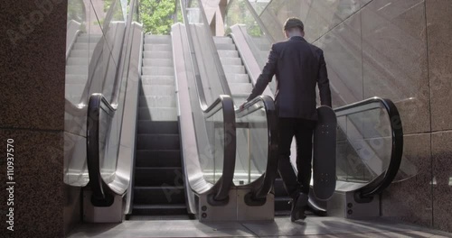 Young man in business suit carries a skateboard while going up an escalator in urban location. Slow motion wide shot recorded at 60fps.