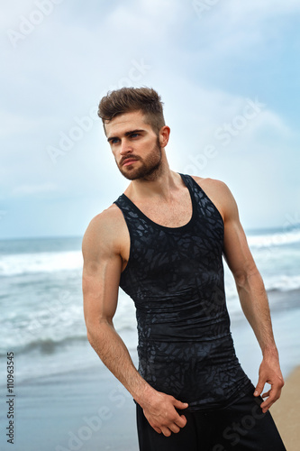 Portrait Of Healthy Handsome Athletic Man With Fit Muscular Body In Summer Sportswear Standing On Ocean Or Sea Beach. Sporty Male Resting After Exercising And Running Outdoor. Sports  Fitness Concept